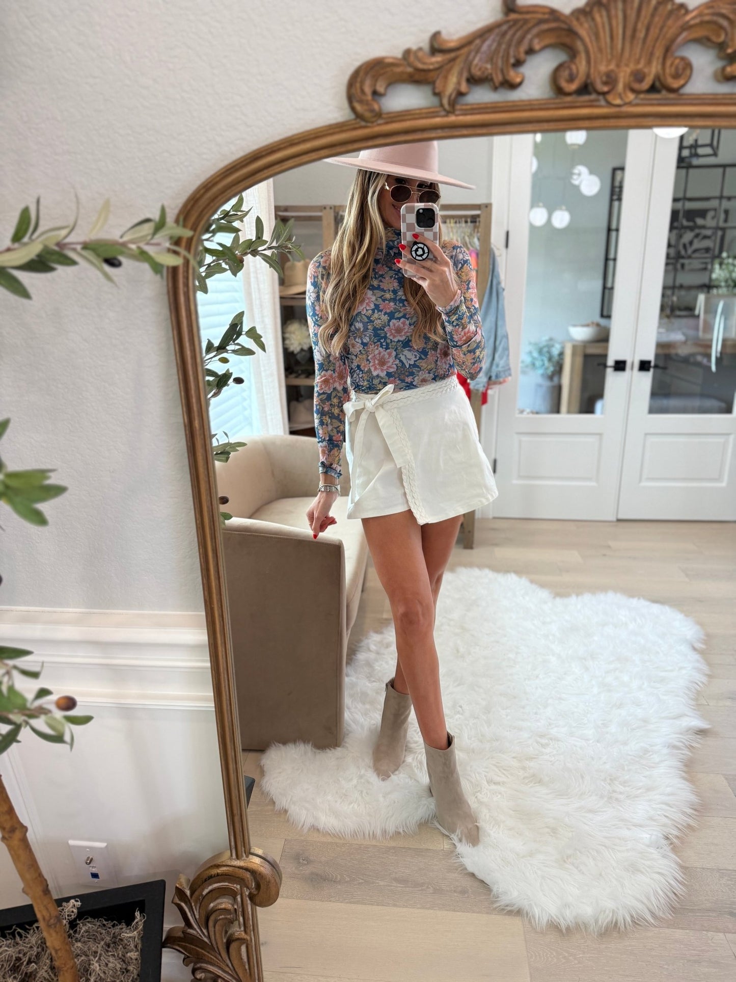 Woman takes a mirror selfie in a Blue Floral Mesh Top, white shorts, beige boots, and a pink hat in a modern room.