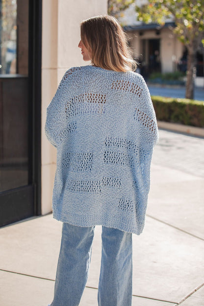 A woman in a Blue Open Front Popcorn Knit Cardigan and jeans stands outside, facing away on a sunny day.
