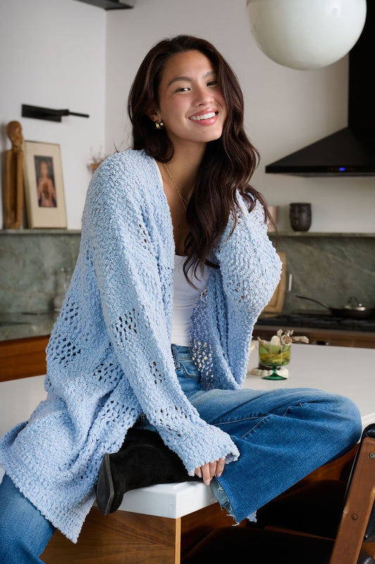 A smiling woman in a Blue Open Front Popcorn Knit Cardigan and jeans sits on a kitchen counter with one leg crossed.