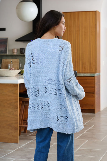 A woman with long dark hair wears a Blue Open Front Popcorn Knit Cardigan and blue jeans in a modern kitchen.
