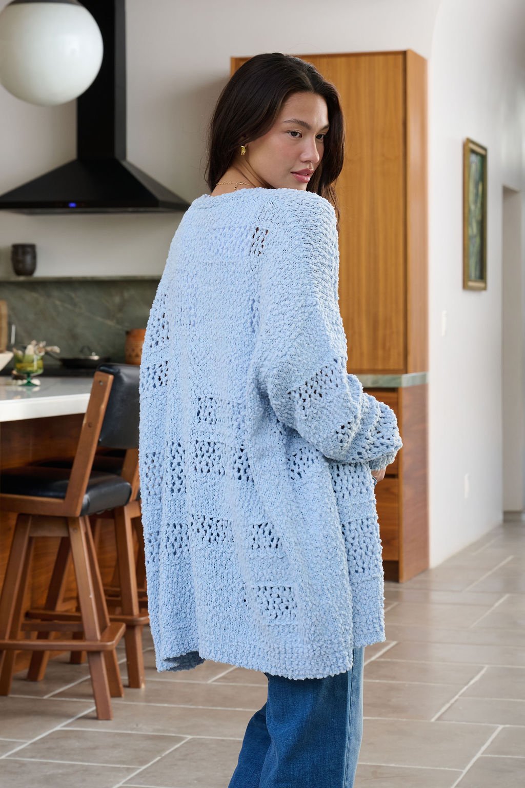 A woman with long dark hair wears a Blue Open Front Popcorn Knit Cardigan and jeans, standing in a modern kitchen.