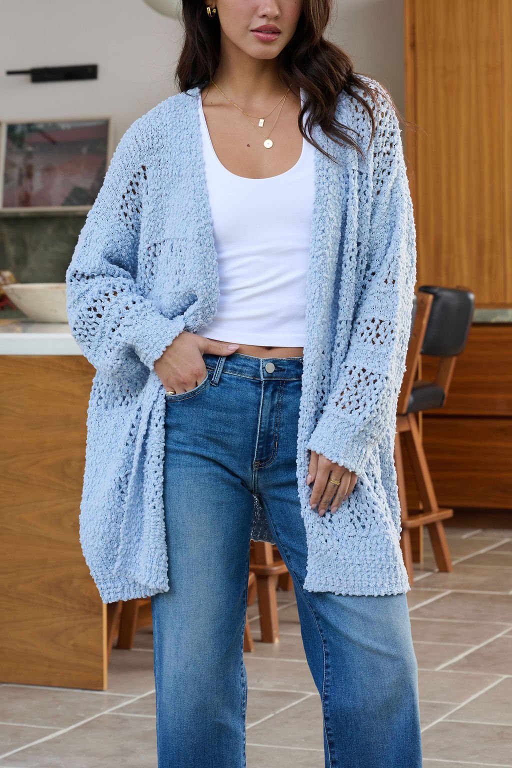 Woman wearing a Blue Open Front Popcorn Knit Cardigan over a white tank top and blue jeans in a modern kitchen.