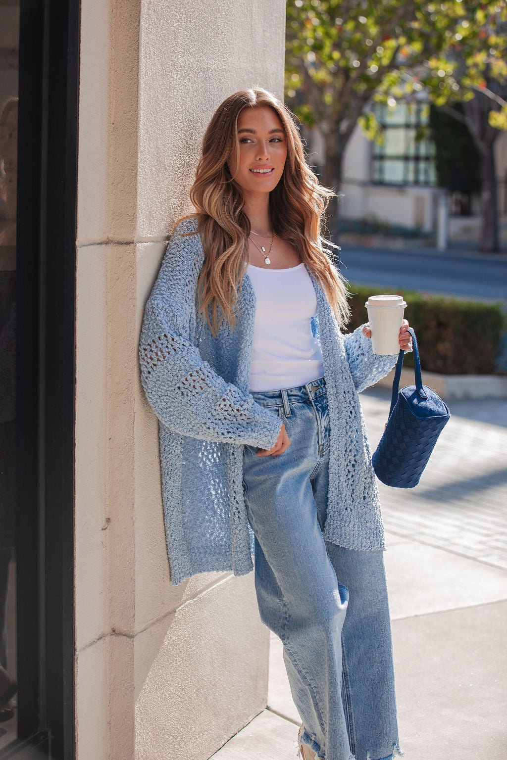 A woman in a Blue Open Front Popcorn Knit Cardigan, white top, and jeans holds a coffee cup and blue woven handbag outdoors in sunlight.
