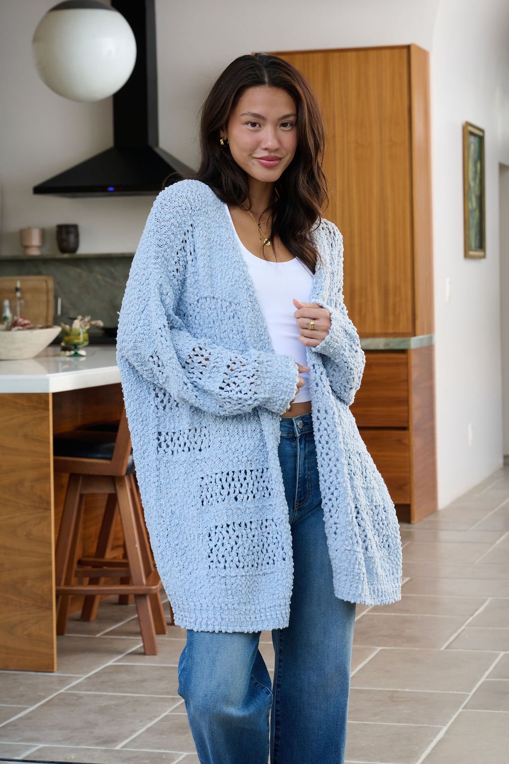 A woman in a Blue Open Front Popcorn Knit Cardigan, white top, and jeans stands in a modern kitchen with wooden cabinets.