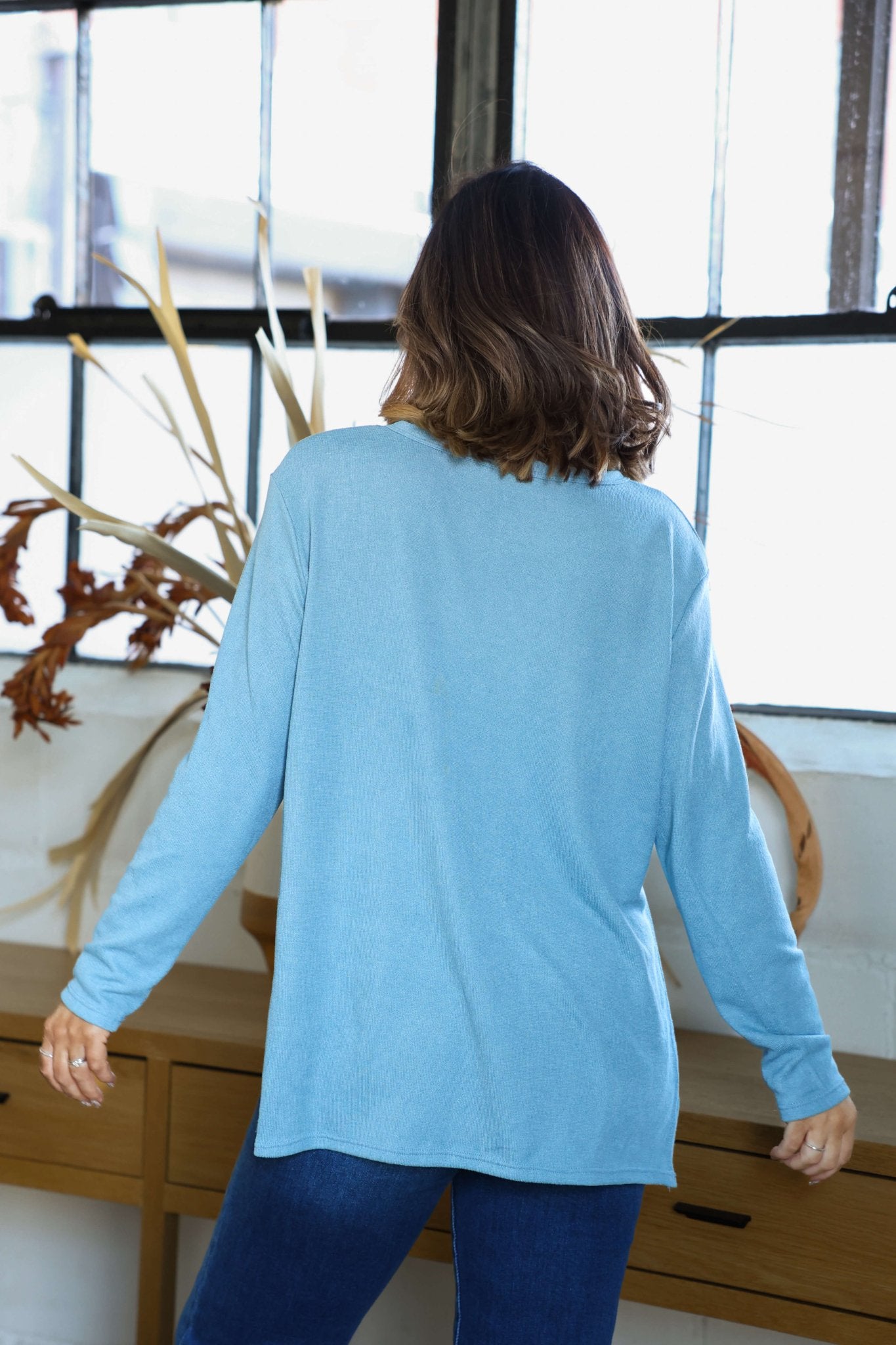 A woman in a Blue Oversized Side Slit Top-FINAL SALE and jeans stands with her back to the camera by a window and wooden dresser.