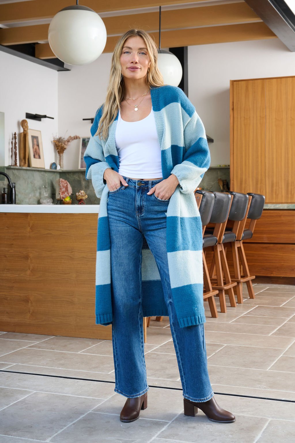 A woman in a Blue Striped Open Front Duster Cardigan and jeans stands in a modern kitchen, ideal for winter wardrobe updates.