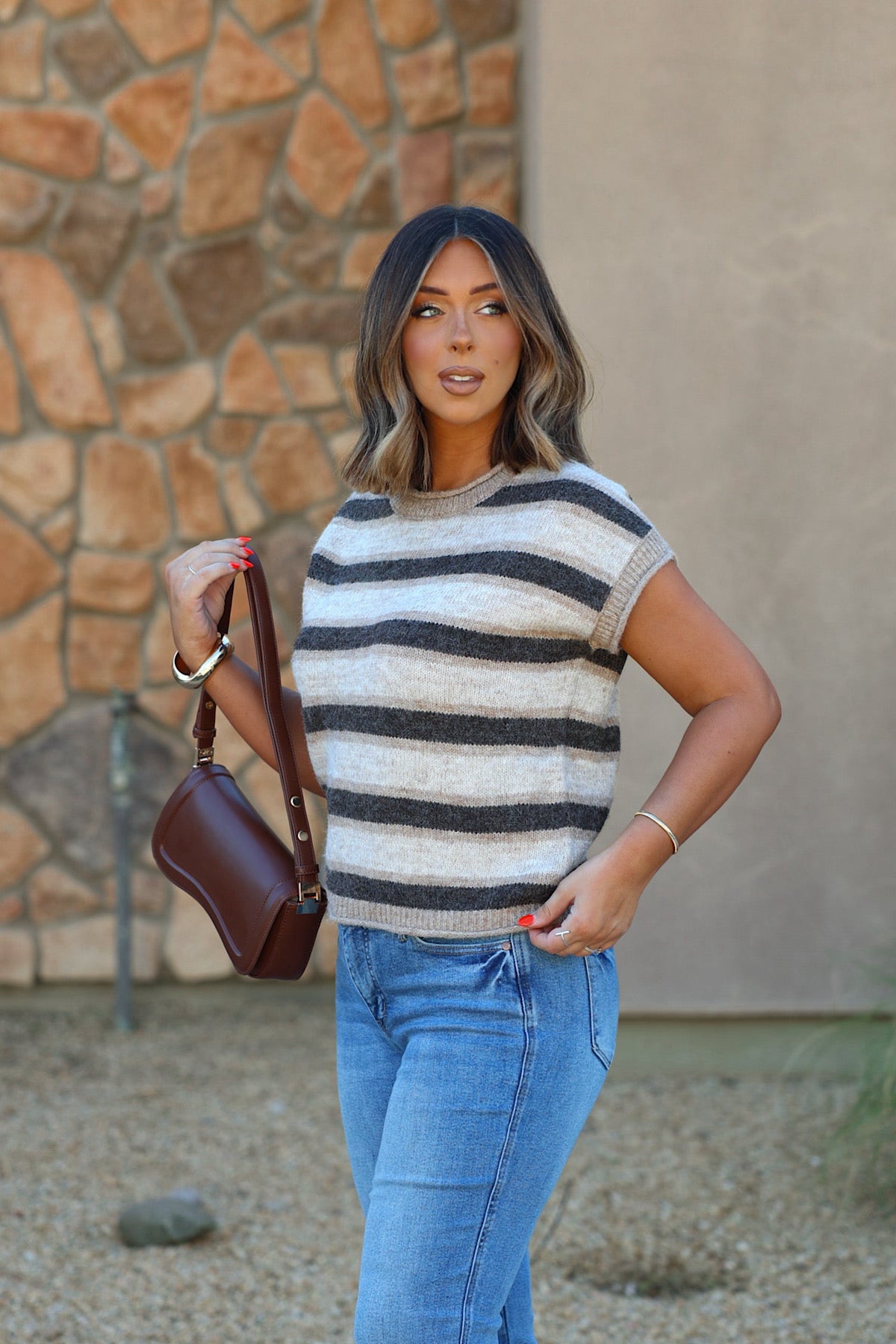 Woman in BLUPEPPER B6FT1055 striped sleeveless sweater, blue jeans, holding a brown bag by a stone wall outdoors.