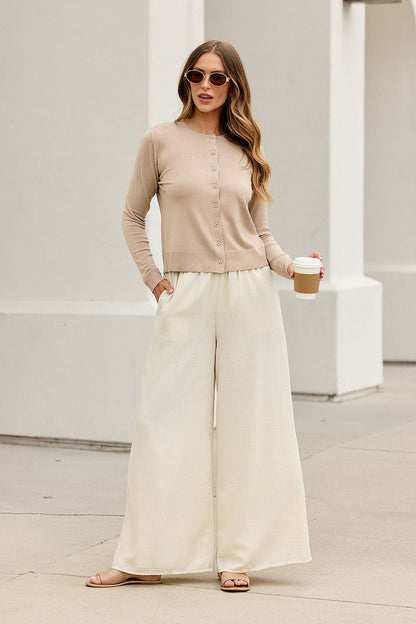 Woman in sunglasses, Boardroom Button Down Sweater - Tan, cream wide-leg pants, and sandals holds coffee—effortless workwear chic.