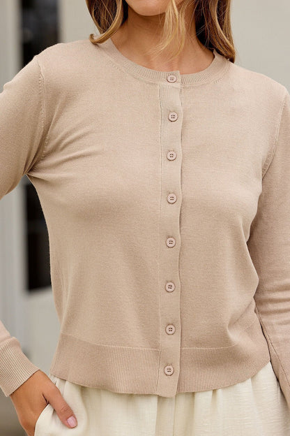 Woman models the Boardroom Button Down Sweater - Tan, paired with a cream skirt, confidently posing indoors with hand on hip.