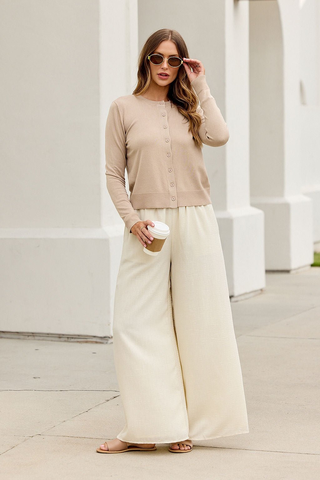 Woman in sunglasses, Boardroom Button Down Sweater - Tan, and cream pants—chic city workwear by a white building.