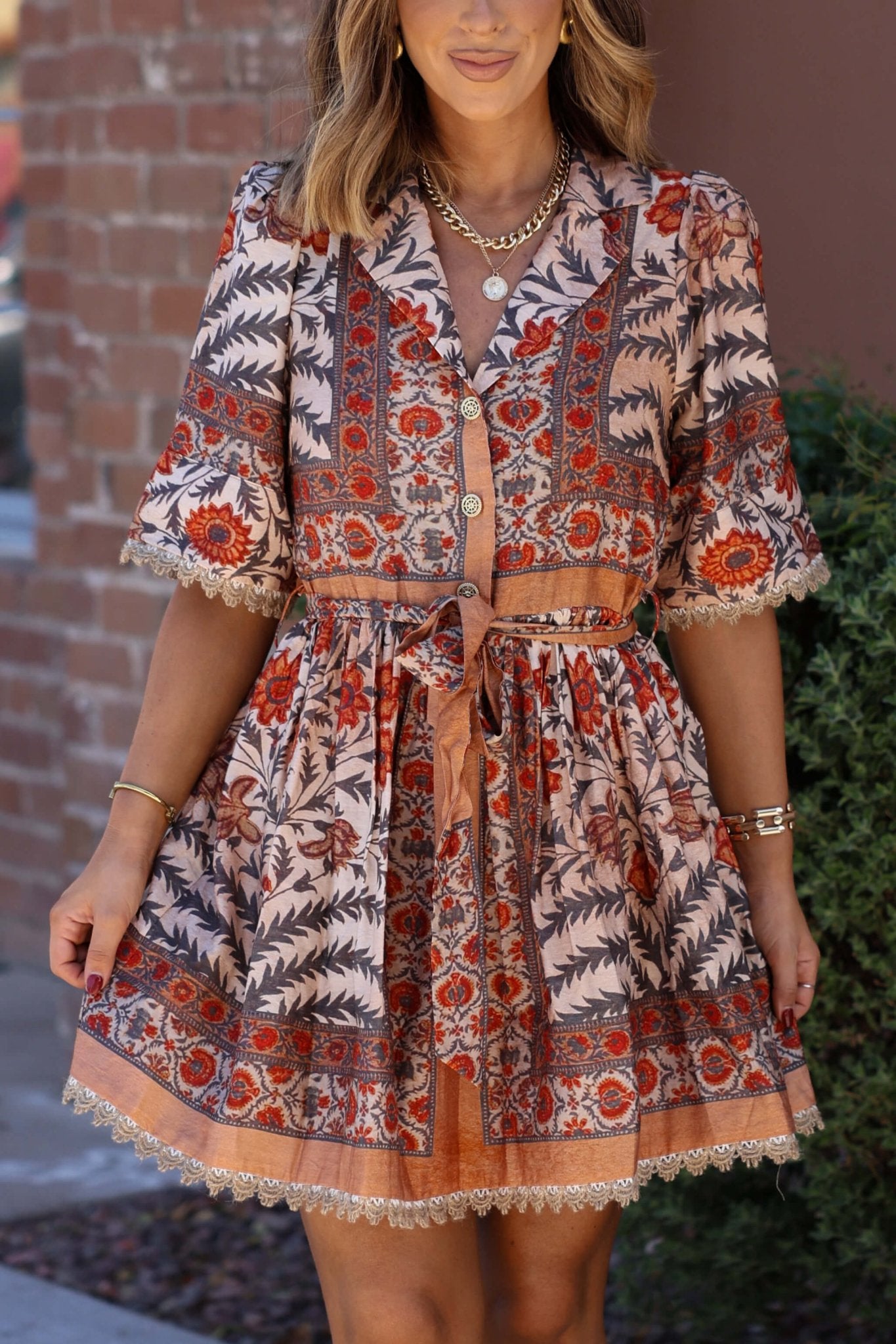 A woman in the Botanical Charcoal Print Belted Mini Dress and gold jewelry stands by a brick wall—ideal for your fall wardrobe.
