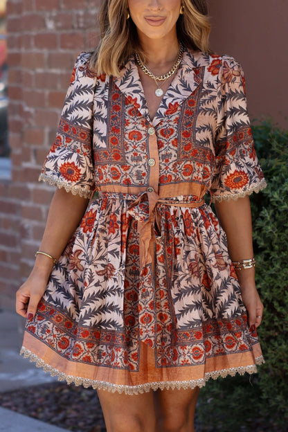 A woman in the Botanical Charcoal Print Belted Mini Dress and gold jewelry stands by a brick wall—ideal for your fall wardrobe.