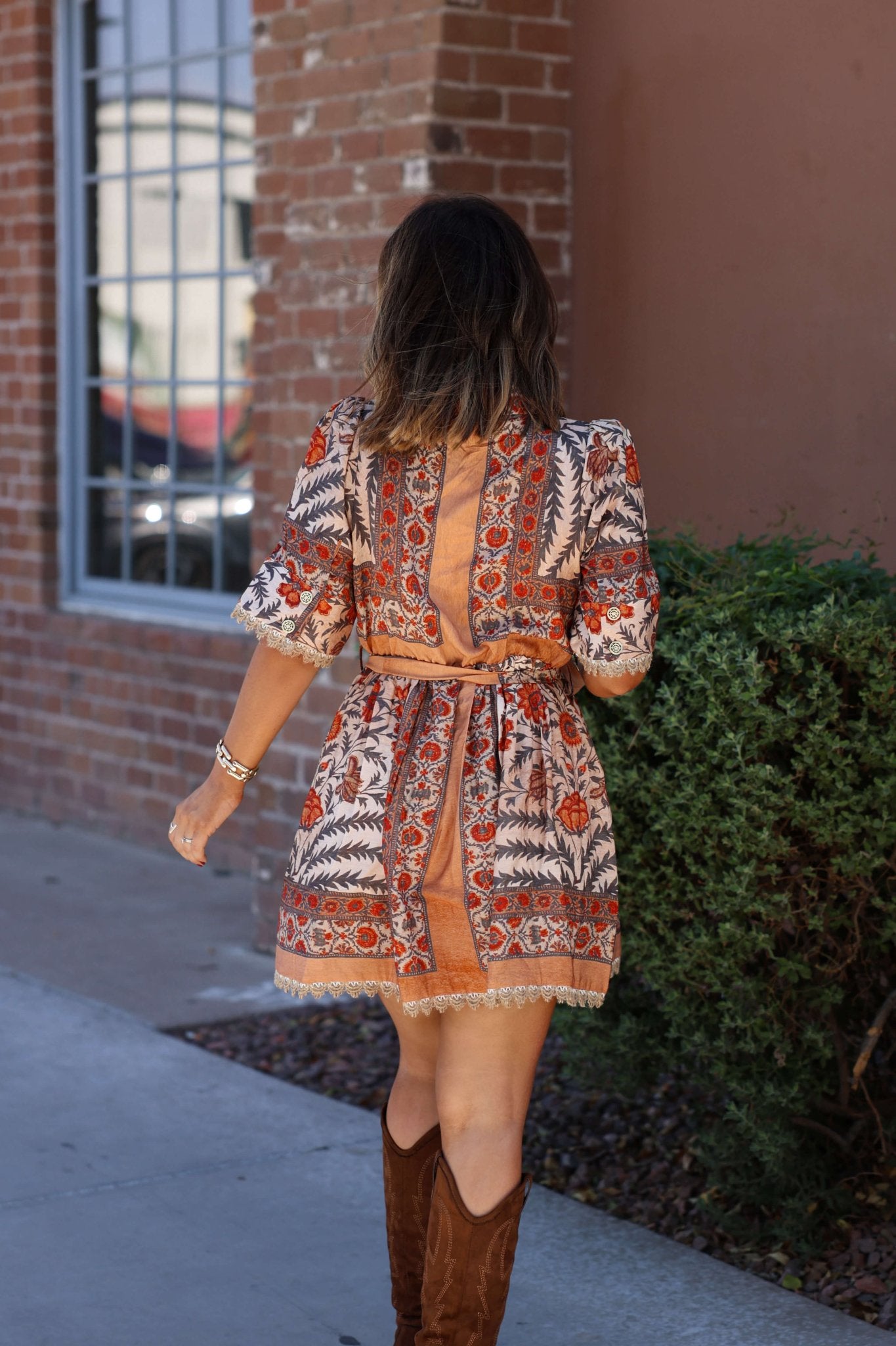 A woman in a Botanical Charcoal Print Belted Mini Dress and brown boots walks on a sidewalk by a brick building with greenery.
