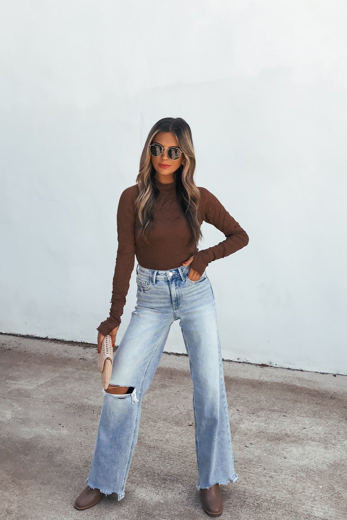 Woman in sunglasses wears the Bramble Lace Top - Brown, ripped jeans, and boots, posing with a clutch against a white wall.