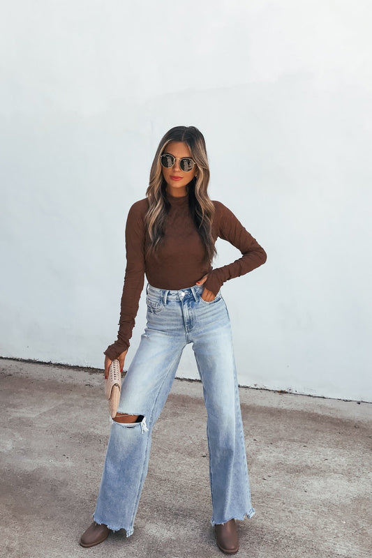 Woman in sunglasses wears the Bramble Lace Top - Brown, ripped jeans, and boots, posing with a clutch against a white wall.