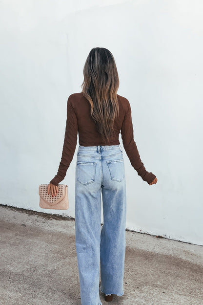 Woman with long wavy hair wears a Bramble Lace Top - Brown and light blue jeans, holding a small pink purse by a white wall.