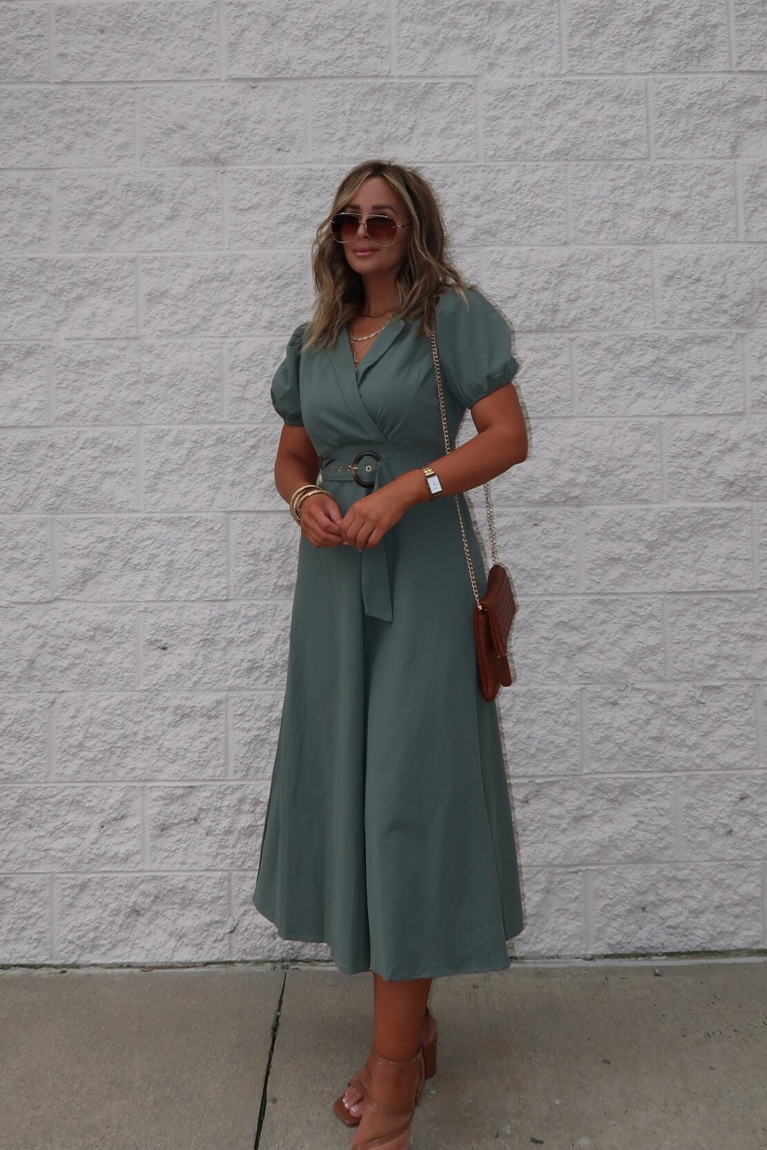 Woman in the Breland Olive Belted Midi Dress stands in heels and sunglasses by a textured white brick wall.