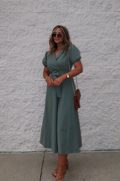 Woman in the Breland Olive Belted Midi Dress stands in heels and sunglasses by a textured white brick wall.