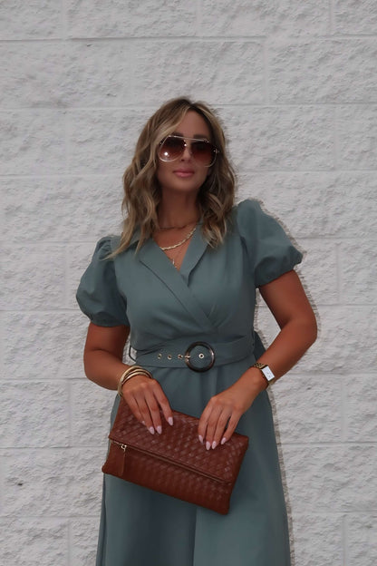 Woman in the Breland Olive Belted Midi Dress stands with sunglasses and a brown clutch against a light textured wall.
