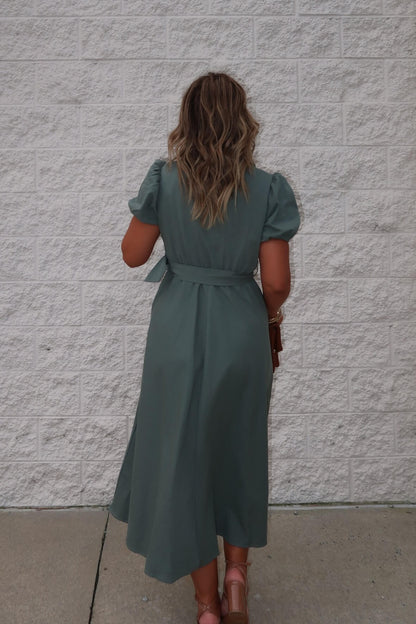 A woman with wavy hair stands by a white brick wall, wearing the Breland Olive Belted Midi Dress - FINAL SALE.