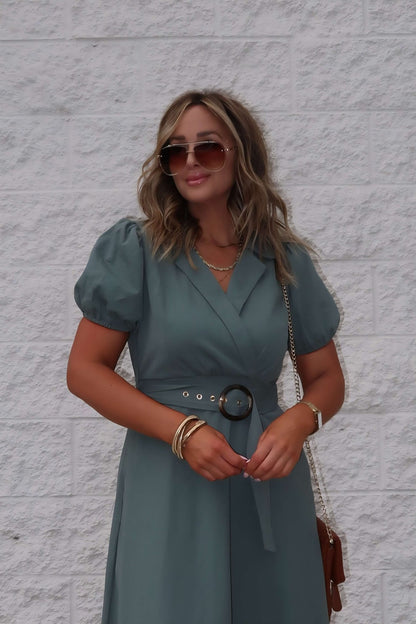Woman in the Breland Olive Belted Midi Dress, sunglasses, gold jewelry, and brown purse stands by a white textured wall.