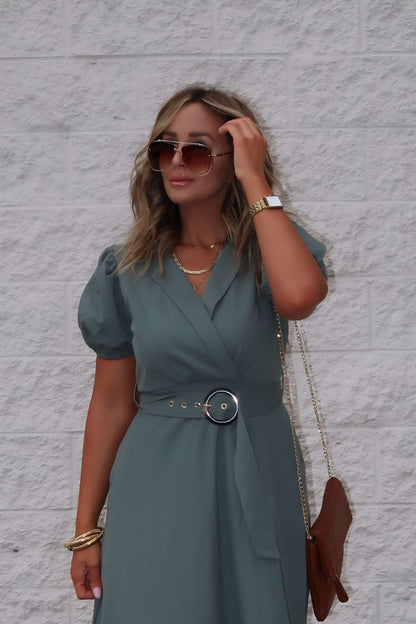 Woman in sunglasses wears the Breland Olive Belted Midi Dress, standing by a white brick wall with a brown purse.
