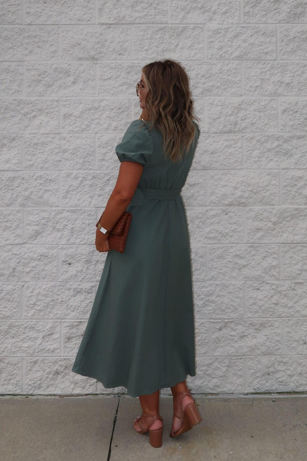 A woman wears the Breland Olive Belted Midi Dress, styled with blush heels and a brown clutch, standing by a white textured wall.
