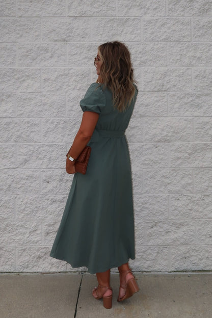 A woman wears the Breland Olive Belted Midi Dress, styled with blush heels and a brown clutch, standing by a white textured wall.