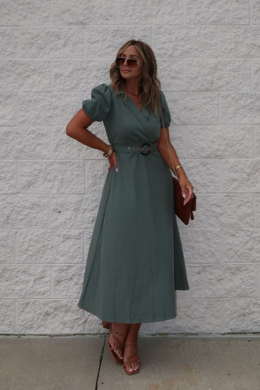 Woman in the Breland Olive Belted Midi Dress, brown sandals, and sunglasses holds a brown clutch by a white brick wall.