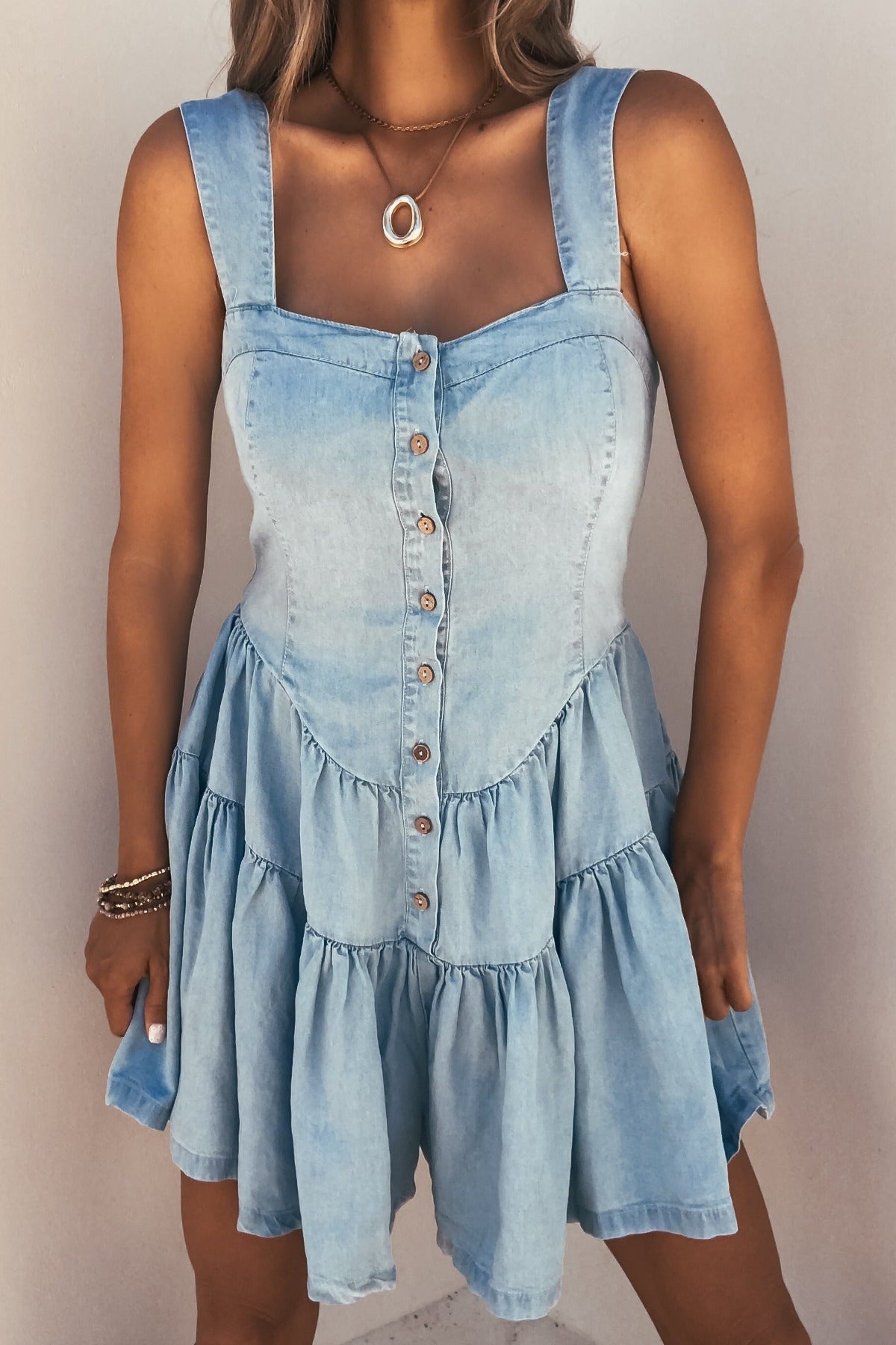 Woman in the Briar Chambray Tiered Romper—sleeveless, light blue with front buttons and ruffle skirt, styled with jewelry.