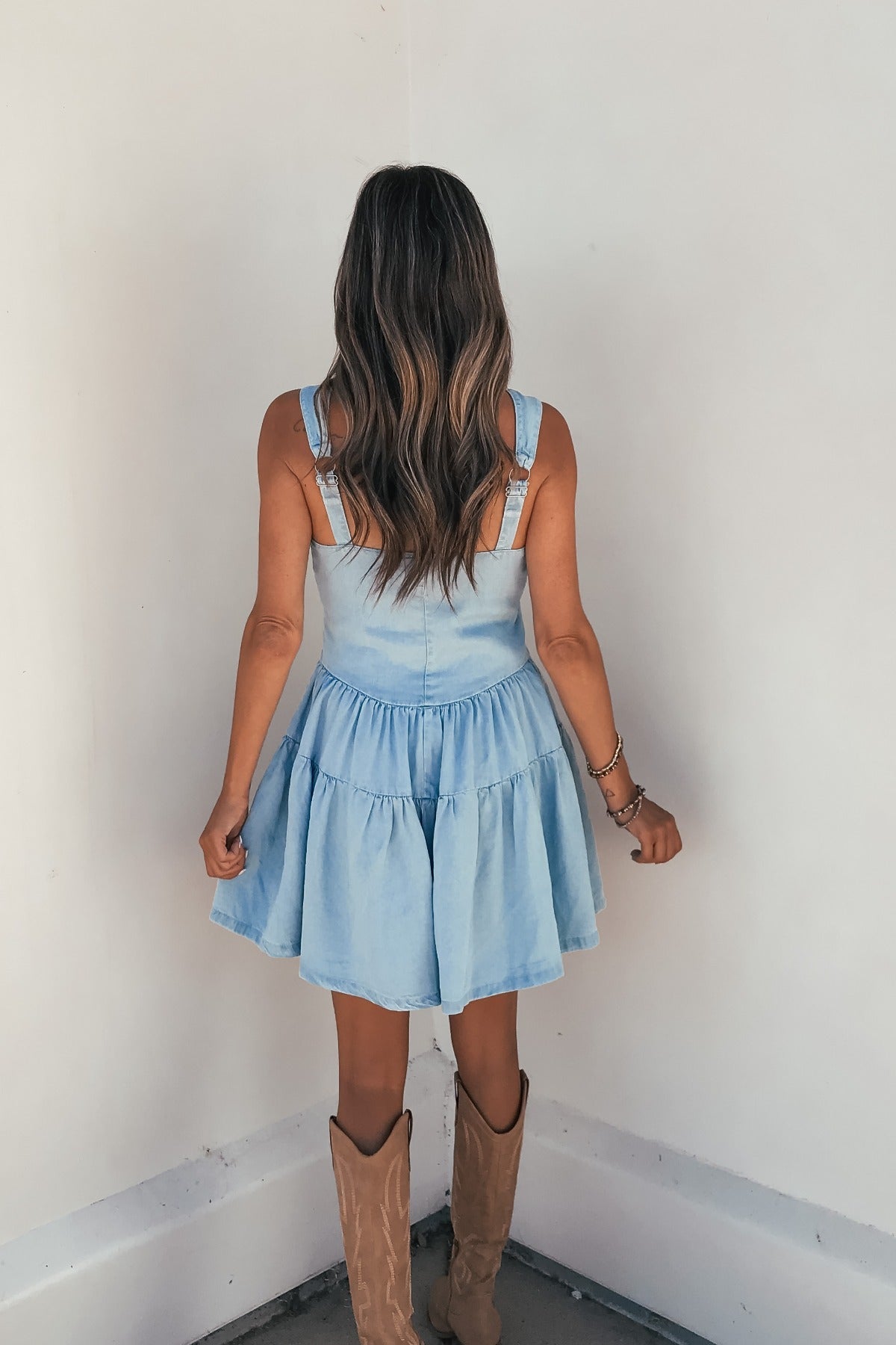 A woman with wavy hair models the Briar Chambray Tiered Romper, standing with her back to the camera facing a white wall.