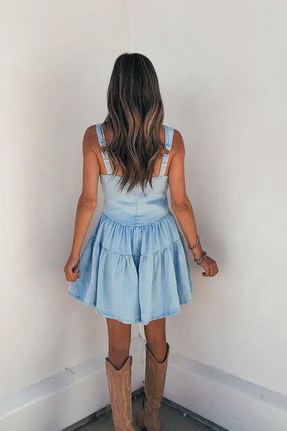 A woman with wavy hair models the Briar Chambray Tiered Romper, standing with her back to the camera facing a white wall.