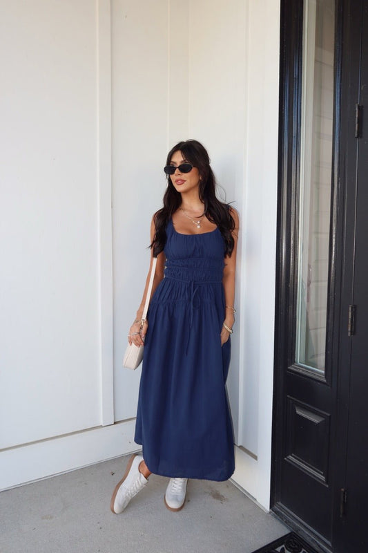A woman in a Bridget Navy Smocked Midi Dress with white sneakers, sunglasses, and a beige handbag stands by a black door.