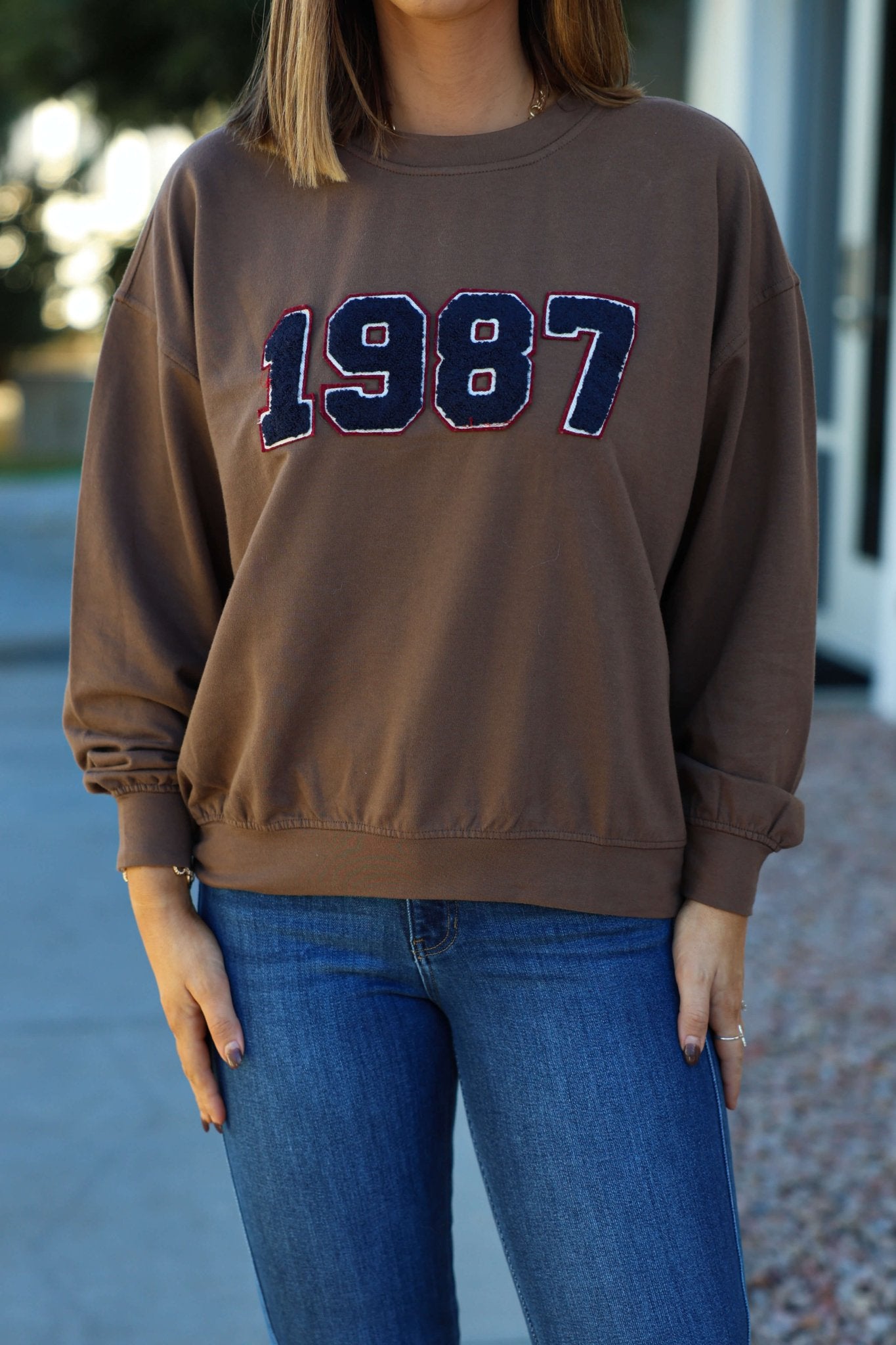 A woman outdoors in a Brown 1987 Long Sleeve Pullover and blue jeans.