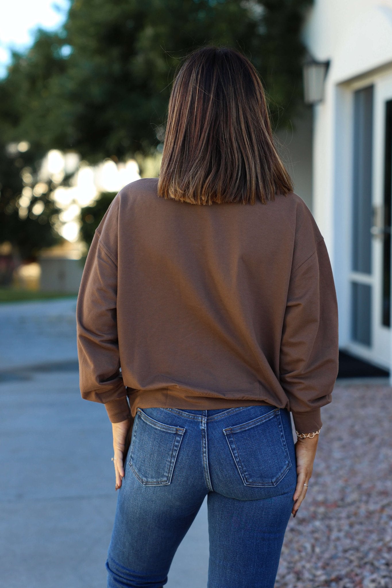 A woman stands outdoors facing away, wearing the Brown 1987 Long Sleeve Pullover with blue jeans and shoulder-length brown hair.