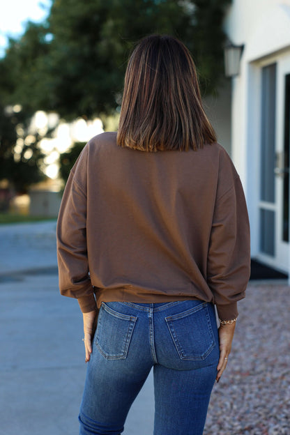A woman stands outdoors facing away, wearing the Brown 1987 Long Sleeve Pullover with blue jeans and shoulder-length brown hair.