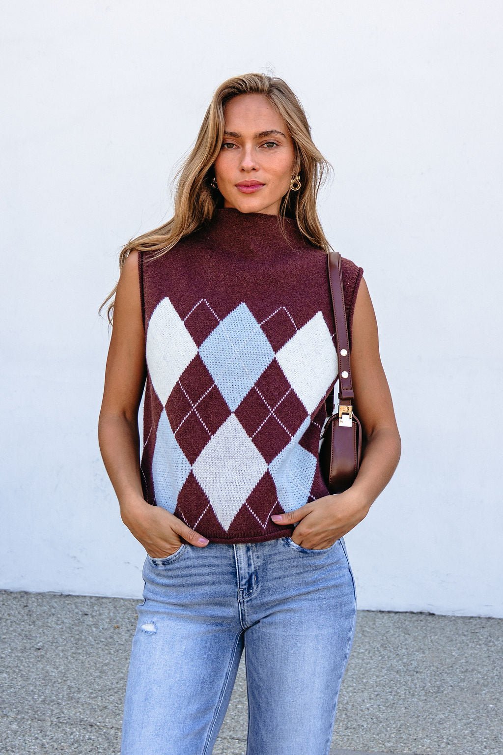 Woman wears a brown argyle sweater vest with light blue jeans, standing by a white wall and holding a brown bag.