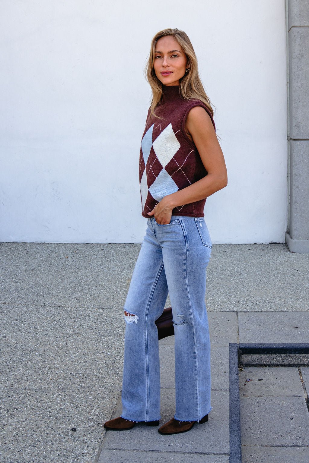 Smiling woman in a Brown Argyle Sweater Vest and light blue jeans stands on pavement outdoors.