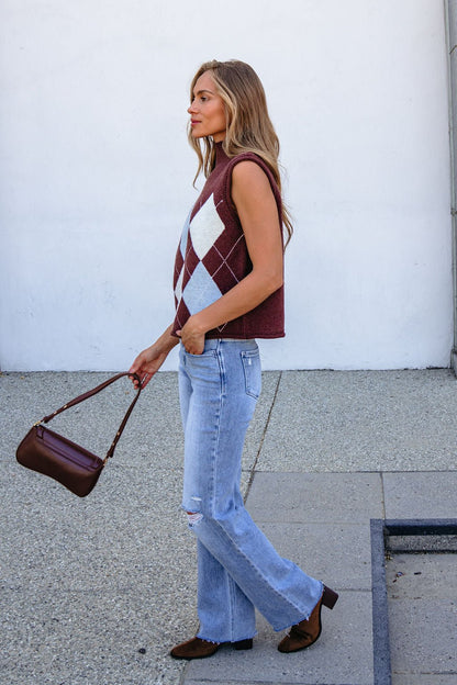 Woman in a Brown Argyle Sweater Vest, relaxed fit, with light blue jeans and brown shoes holds a purse outside by a plain wall.