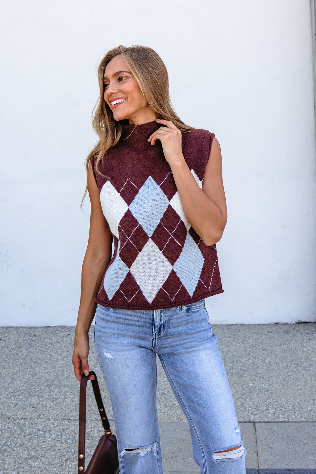 Woman wearing Brown Argyle Sweater Vest and ripped jeans stands smiling outdoors, holding a brown handbag.