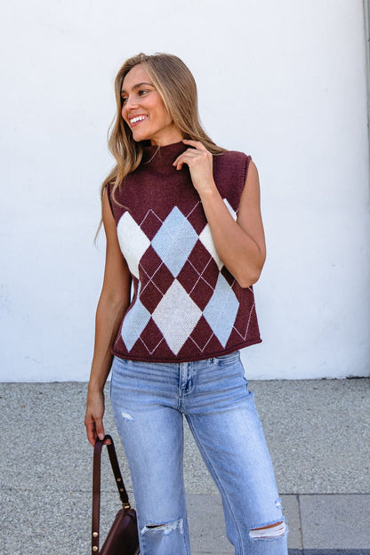 Woman wearing Brown Argyle Sweater Vest and ripped jeans stands smiling outdoors, holding a brown handbag.