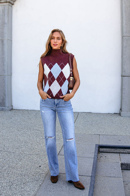 Model wears Brown Argyle Sweater Vest, ripped light blue jeans, and brown shoes outdoors against a neutral backdrop.