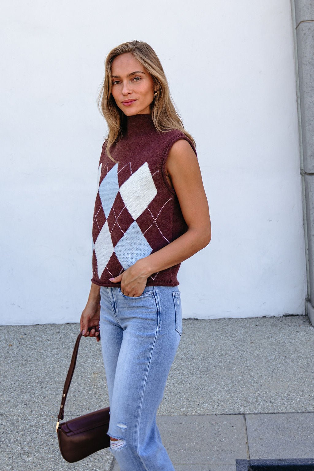 Woman in a Brown Argyle Sweater Vest and light blue jeans stands outside holding a small brown handbag, smiling slightly.