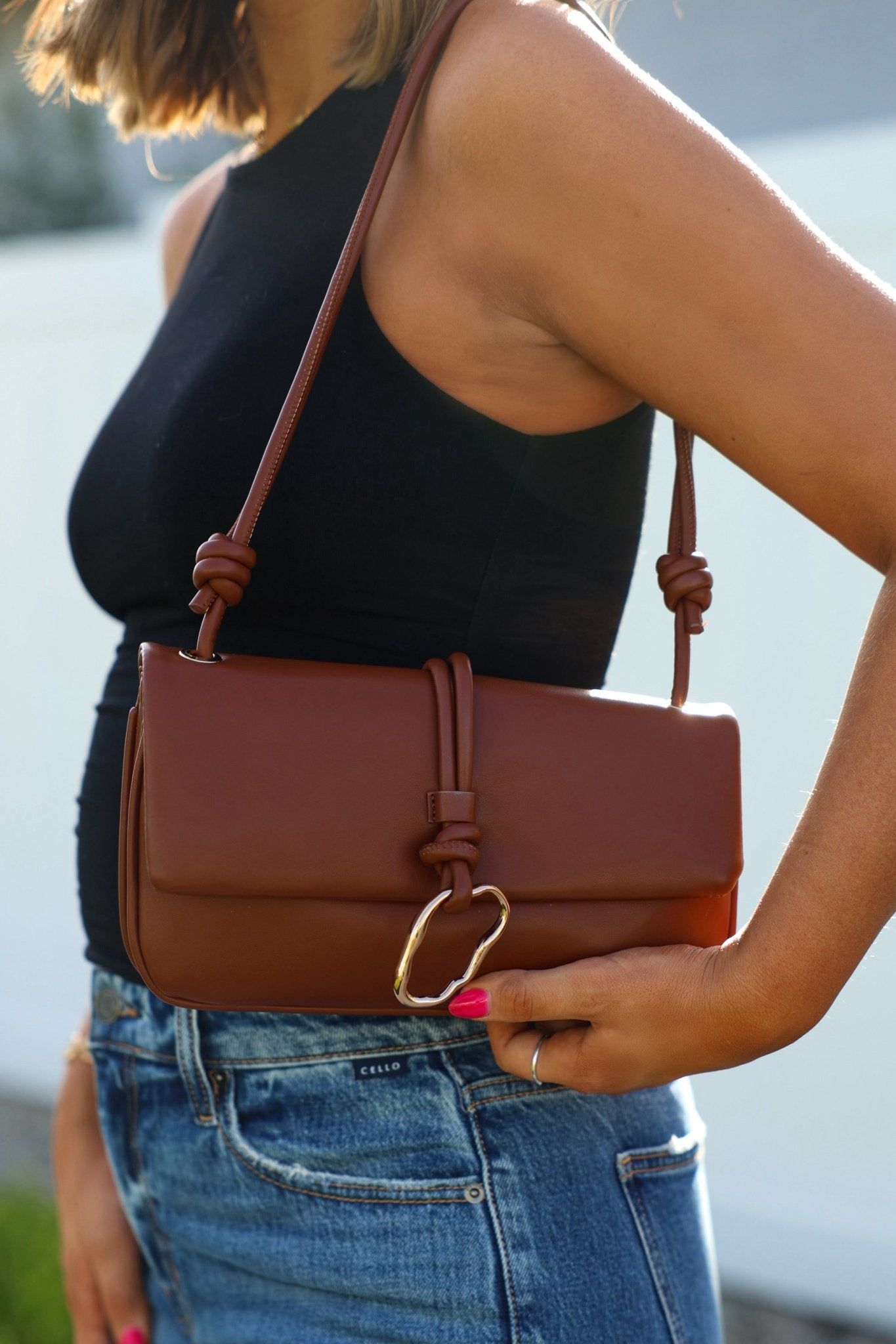 A person in a black sleeveless top and jeans holds the Brown Belina Vegan Leather Baguette Shoulder Bag.