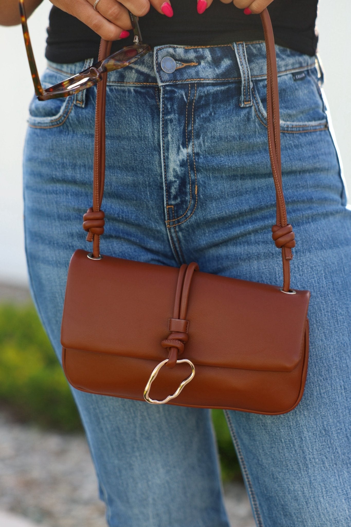 Person in blue jeans holds a Brown Belina Vegan Leather Baguette Shoulder Bag with knot details, gold clasp, and bright pink nails.