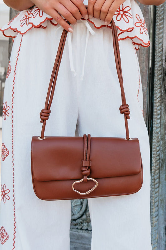 A person in white pants with red embroidery holds the Brown Belina Vegan Leather Baguette Shoulder Bag with gold accents and knotted straps.