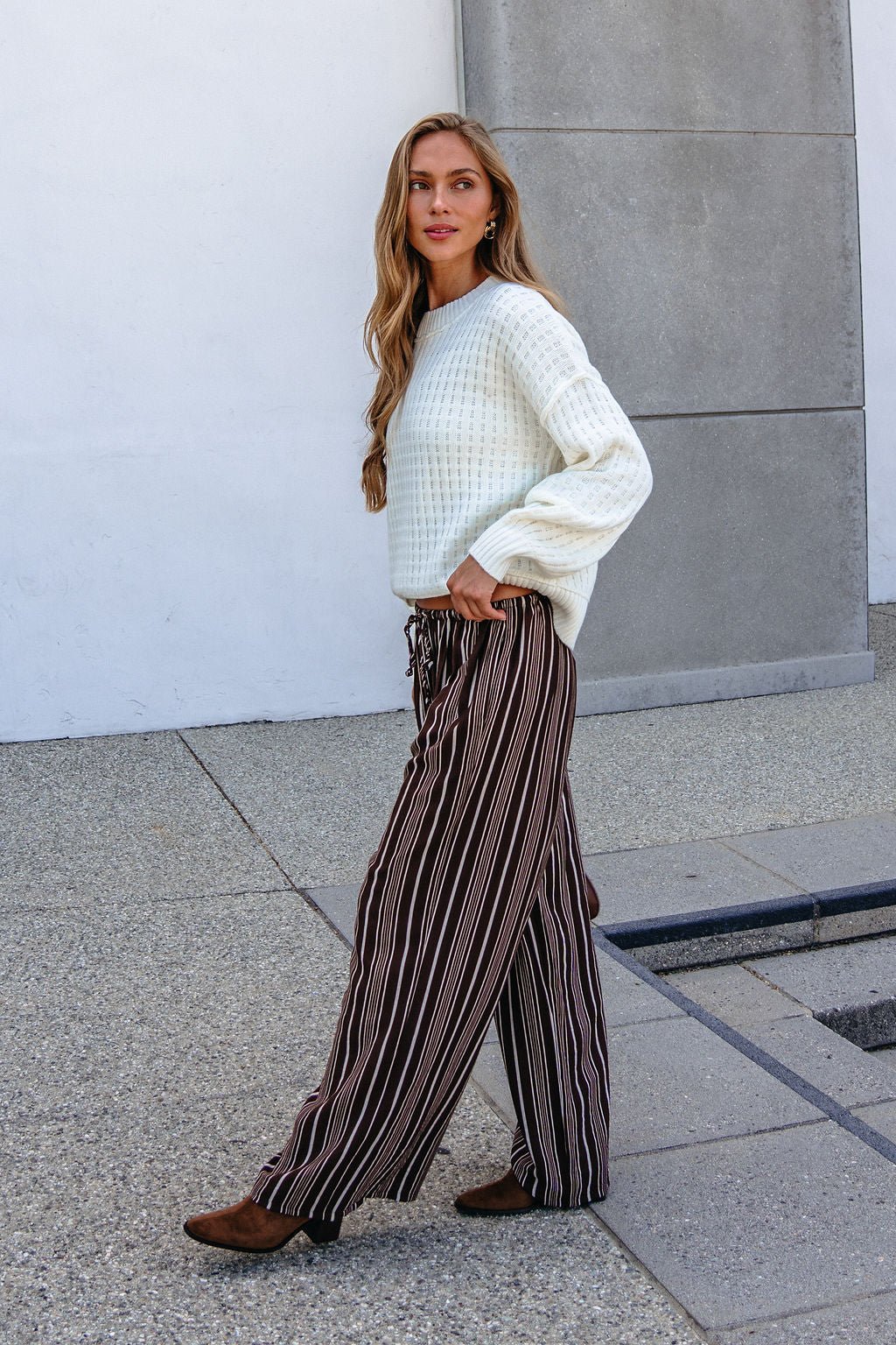 Woman in Brown & Cream Striped Drawstring Pants walks outdoors on a paved path, with a neutral wall in the background.