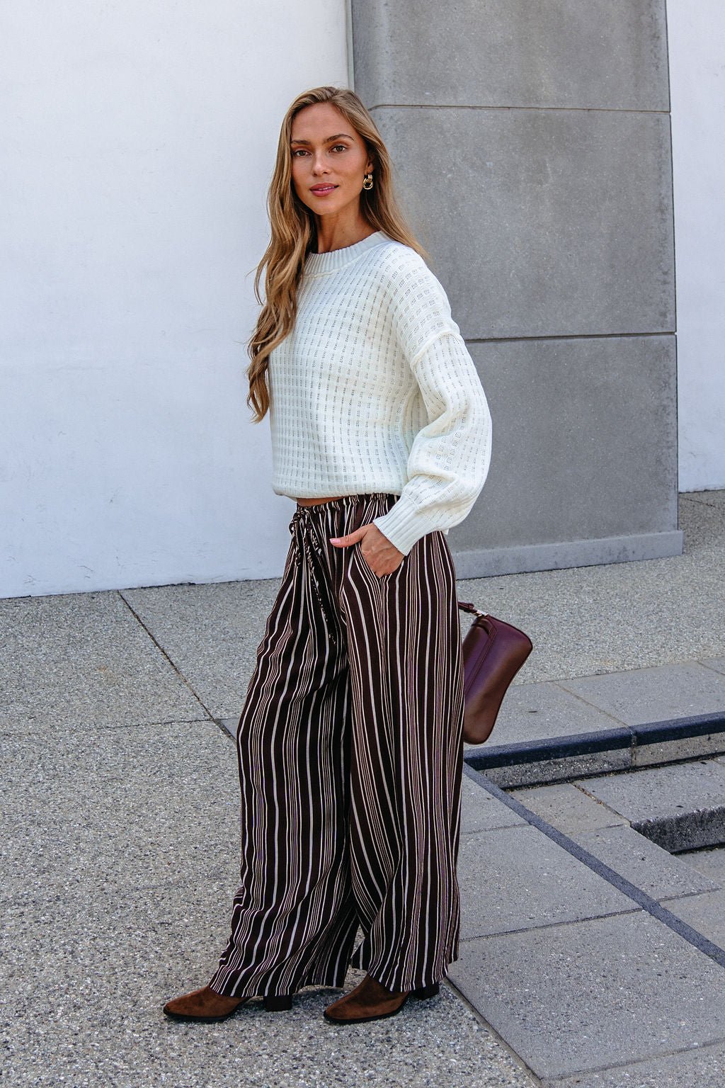 Woman in a white sweater and Brown & Cream Striped Drawstring Pants stands outdoors by a white and gray wall.