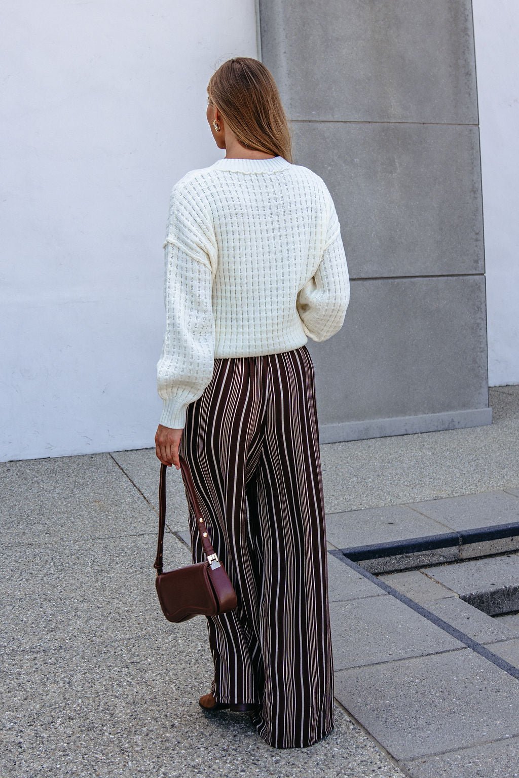 Woman stands outside in a white sweater and Brown & Cream Striped Drawstring Pants, holding a small brown handbag.