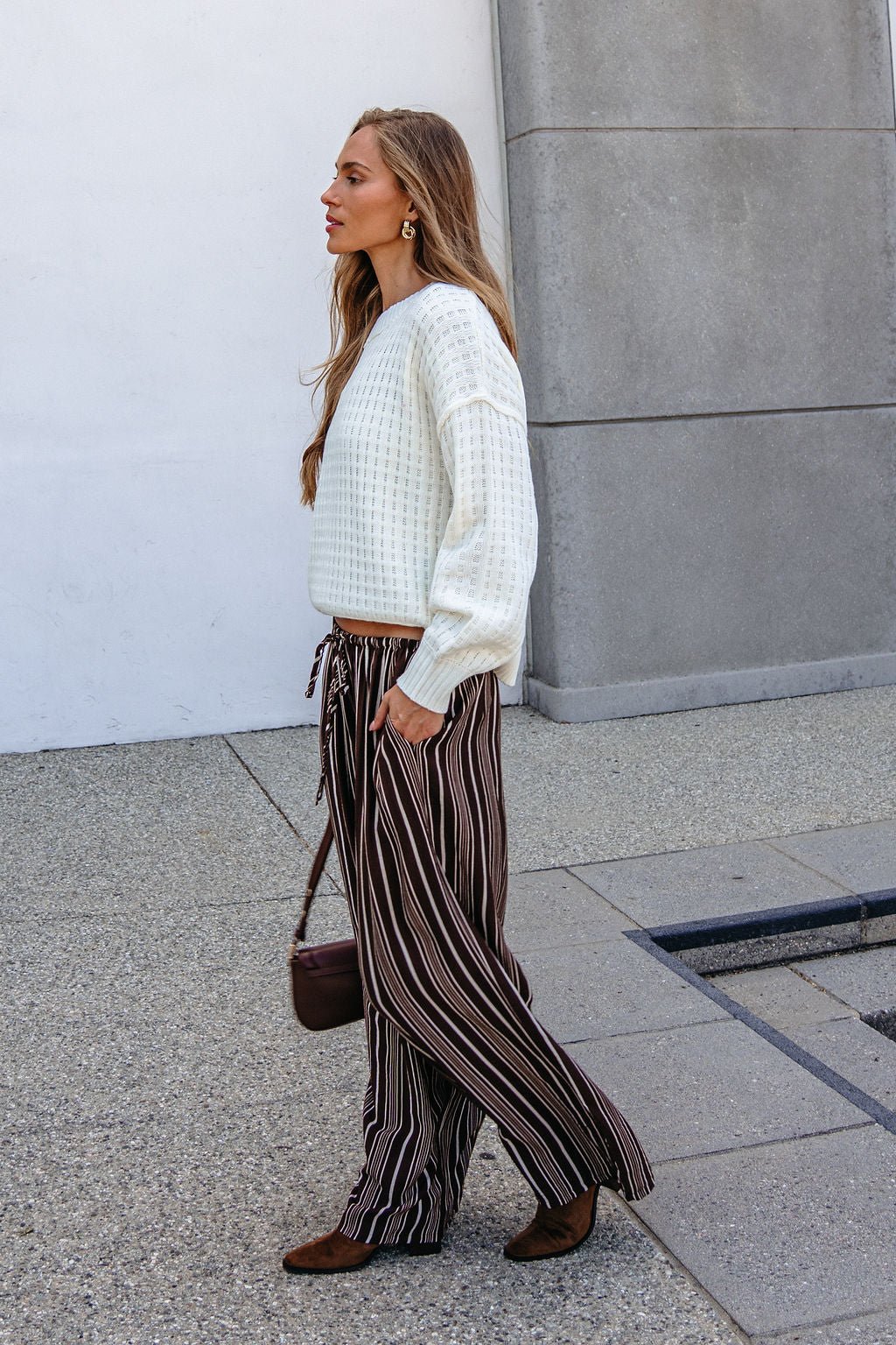 A woman walks outside in a white sweater, Brown & Cream Striped Drawstring Pants, brown shoes, and a matching brown bag.